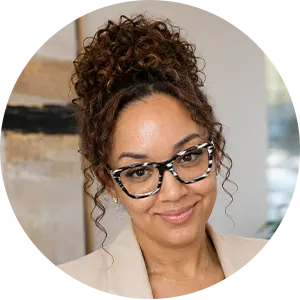 Portrait of a smiling woman with curly hair up, wearing patterned glasses and a beige blazer, indoors.