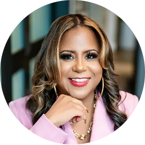 Portrait of a smiling woman with light brown hair in a pink blazer, resting chin on her hand in an office setting.