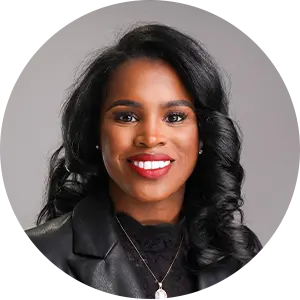 Portrait of a smiling professional woman with dark wavy hair wearing a black blazer against a gray background.